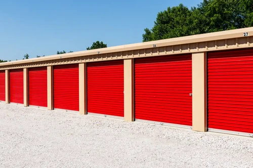 Alt text: Row of outdoor storage units with red roll-up doors under clear blue sky.