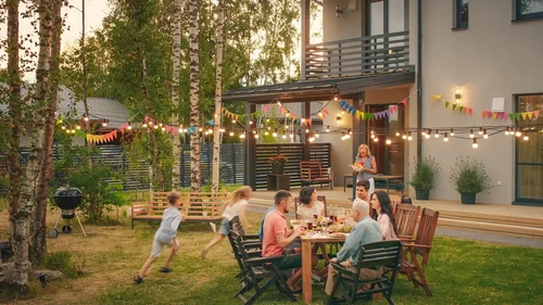 Family and friends gathering for an outdoor meal in a backyard with string lights outside a modern home.
