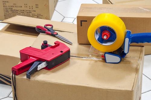 Packing tape dispenser, labeling tool, and scissors on cardboard boxes ready for moving and packing.