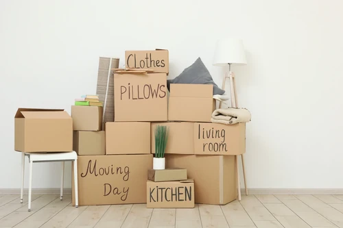 Labeled moving boxes with pillows, books, a lamp, and a blanket stacked in an empty room.