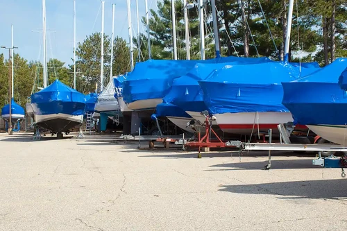 Boats covered with blue tarps stored outdoors on trailers in a storage lot.