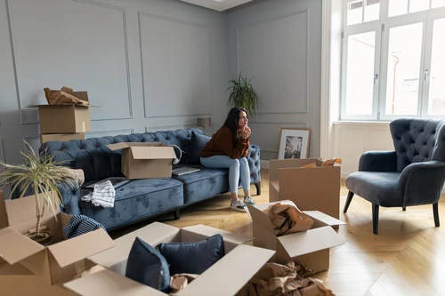 Woman sitting on a couch surrounded by moving boxes in a living room.
