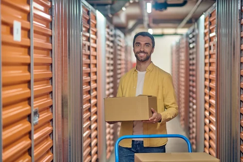 Man holding a box in a hallway of indoor storage units next to a moving cart.