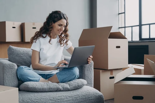 Woman sitting on chair using laptop surrounded by moving boxes at home. 
