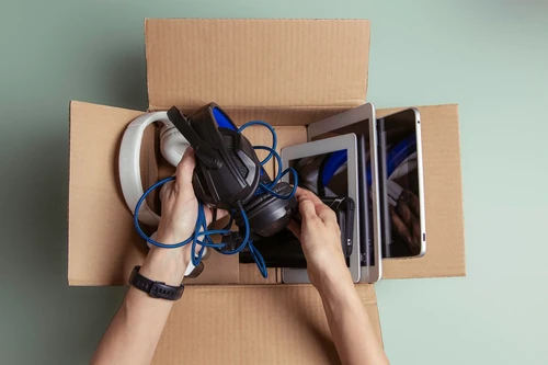 Hands placing electronics and headphones into a cardboard box with tablets and cables.