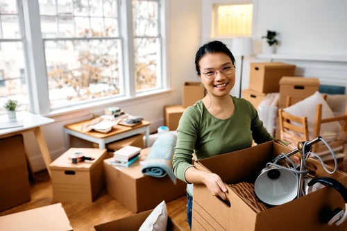 Woman holding box while packing items in home with moving boxes around.