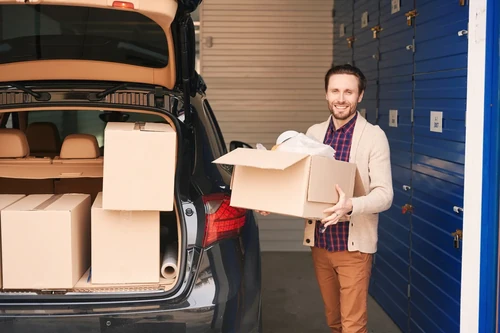 Man holding cardboard box next to open car trunk with stacked boxes near storage units
