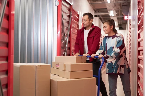 Two people pushing a cart with stacked boxes through an indoor storage hallway with red roll-up doors on both sides.