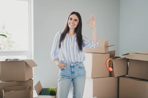 Smiling woman standing with cardboard boxes making OK hand gesture in bright room. 