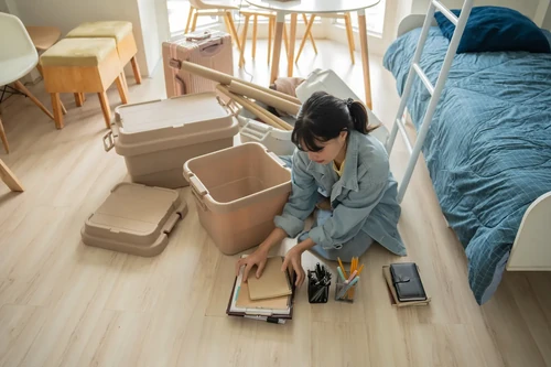 Student sitting on the floor organizing notebooks and packing items into storage bins in a bedroom.
