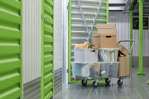 Cart with plastic bins and cardboard boxes in hallway of indoor storage units with green doors.