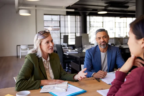 People talking across a desk during an office meeting, with documents and notebooks spread out in a modern workspace.