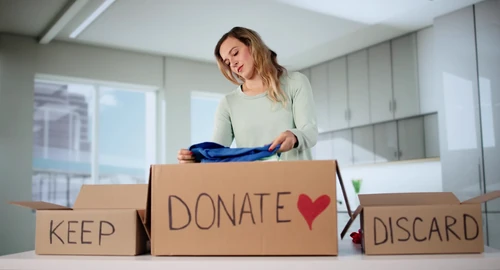 Woman sorting clothes into labeled keep, donate, and discard boxes on a table during a home decluttering process,