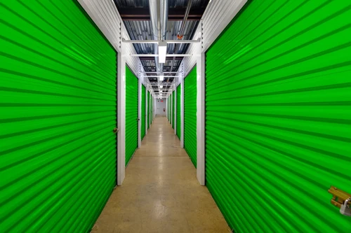Indoor storage hallway with green roll-up doors on both sides, concrete floor, and overhead lights and piping.