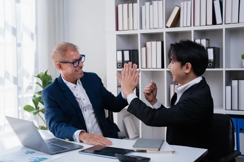 Two businessmen smiling and high-fiving at a desk in an office with laptop, documents, and shelves in the background.