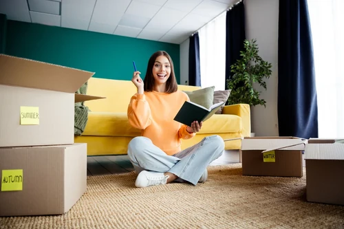 Woman sitting on the floor with labeled moving boxes, holding a notebook and organizing items in a bright living room.