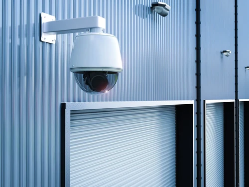 Security camera above storage unit doors on a metal building, monitoring access along a row of roll-up units.