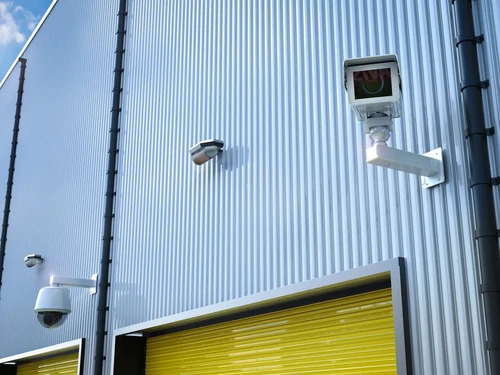 Storage facility exterior with multiple video recording cameras mounted on metal siding above yellow roll-up doors.