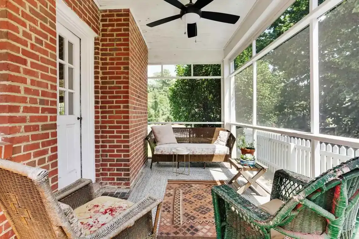 A cozy enclosed porch with wicker furniture, a rug, and plants on a sunny day. 